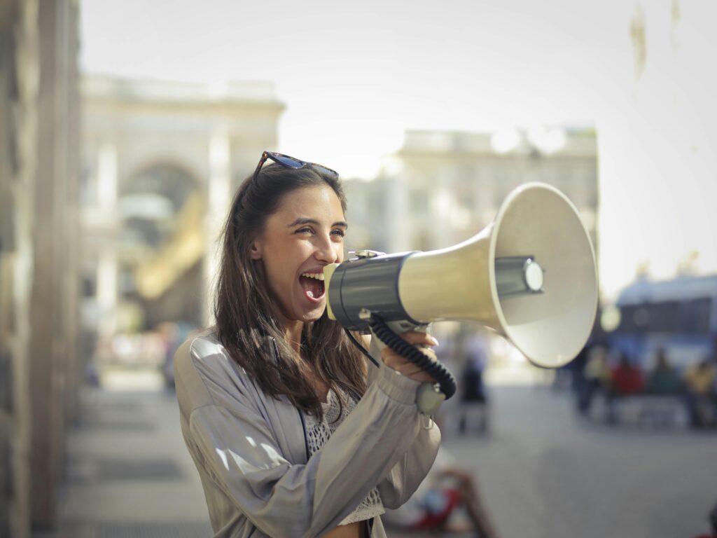 pexels-photo-3761509-3761509 Cheerful young woman in a casual outfit shouting into a megaphone on a sunny day.